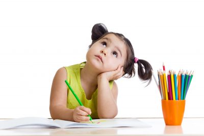 Young girl thinking while writing, with a cup of coloured pencils
